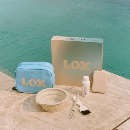 LOX complete kit including a washbag, box, bowl, brush, mirror and bottle on a stone surface with water in the background.